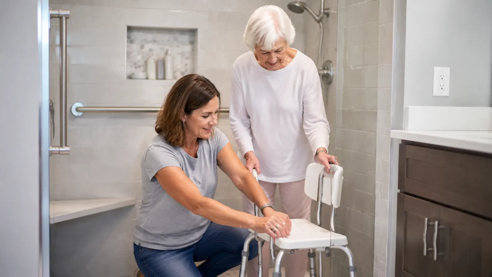 Adult daughter helping her elderly mother adjust a shower bench, both smiling, bright bathroom