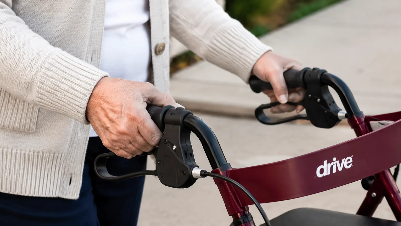 Close-up of hands gripping the Drive Medical rollator handle, brake levers visible, standing on a sidewalk
