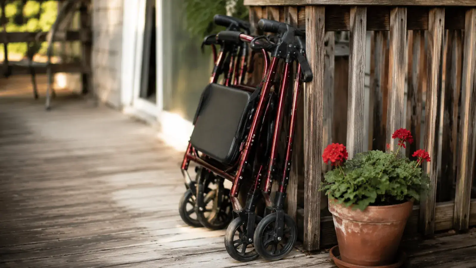 A burgundy rollator walker parked on a front porch next to a potted plant, folded neatly against the railing