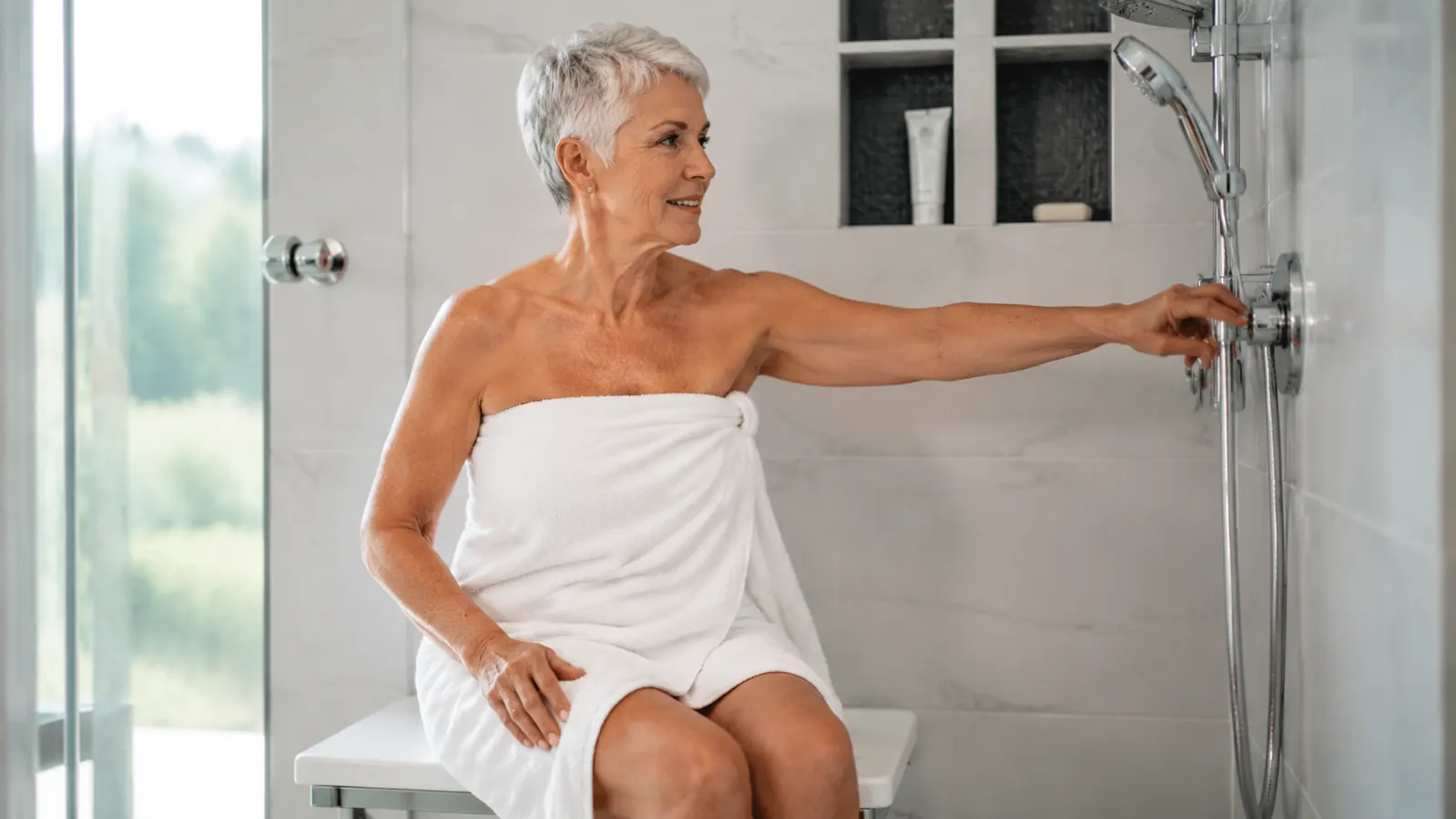 Woman seated on shower bench reaching to adjust water temperature at shower controls within easy reach